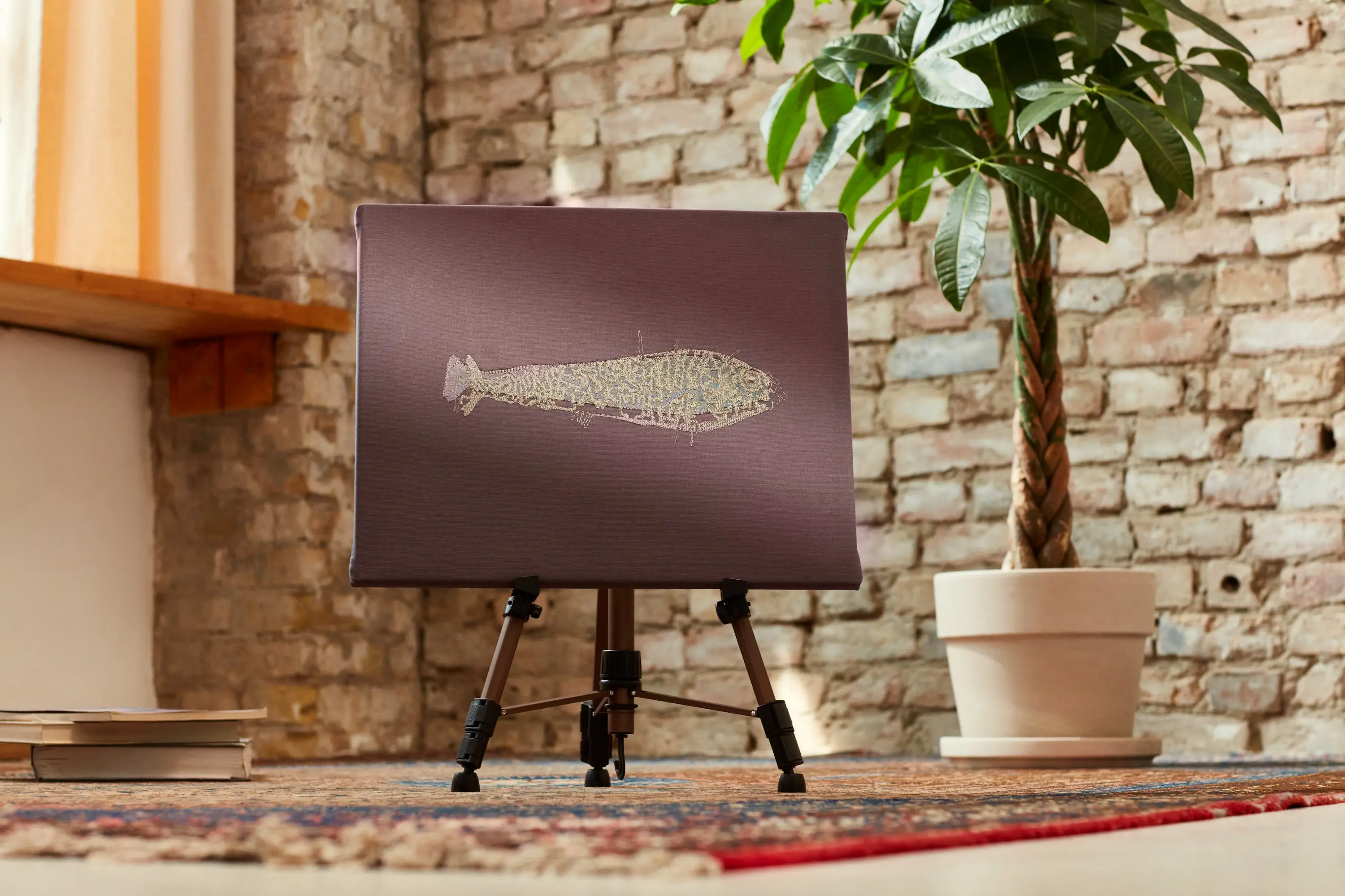 Gesticktes Fischbild in Gold auf dunklem Stoff, auf einer Staffelei vor einer Backsteinwand