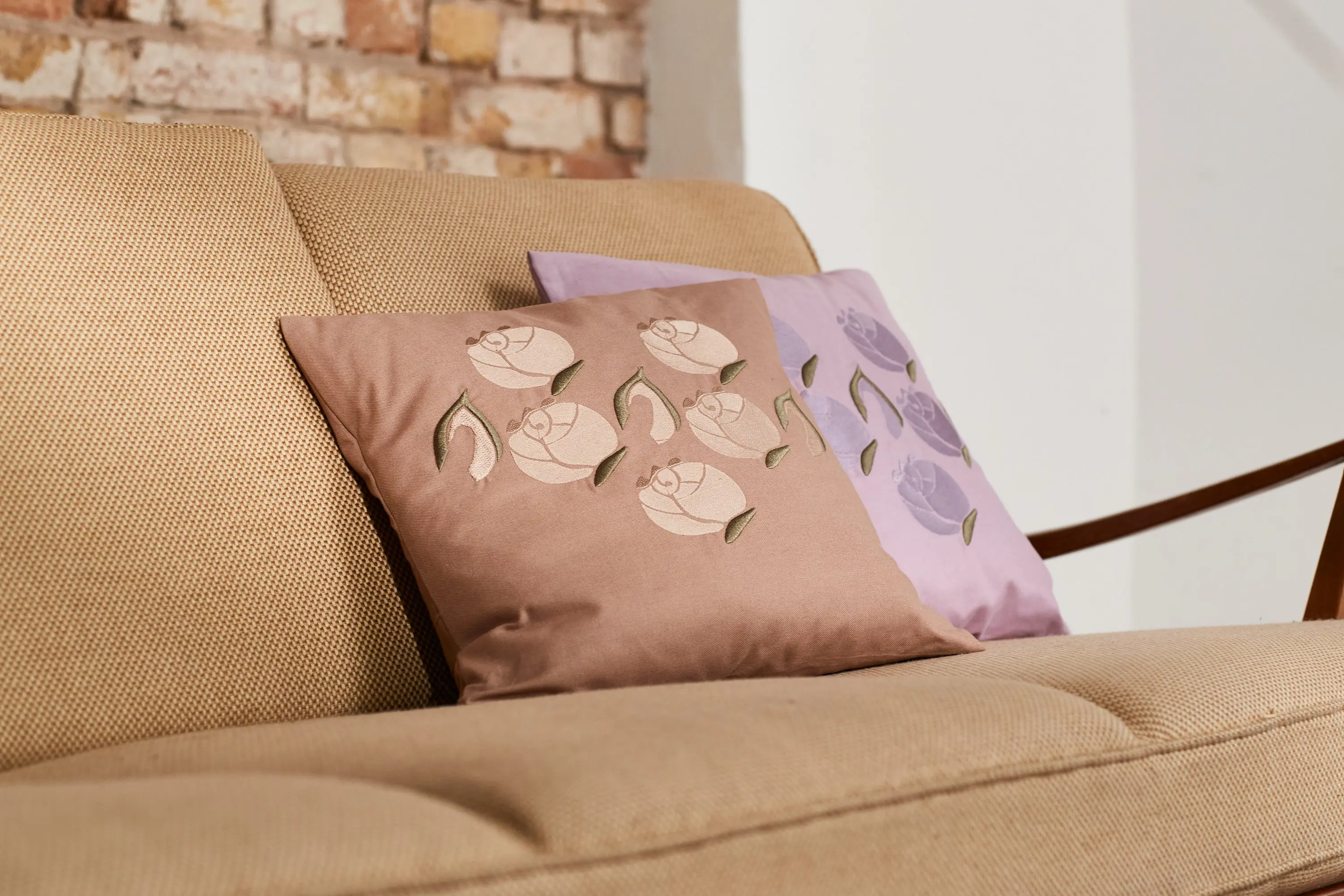 Bestickte Kissen in Rosé und Flieder auf einem Sofa vor einer Backsteinwand