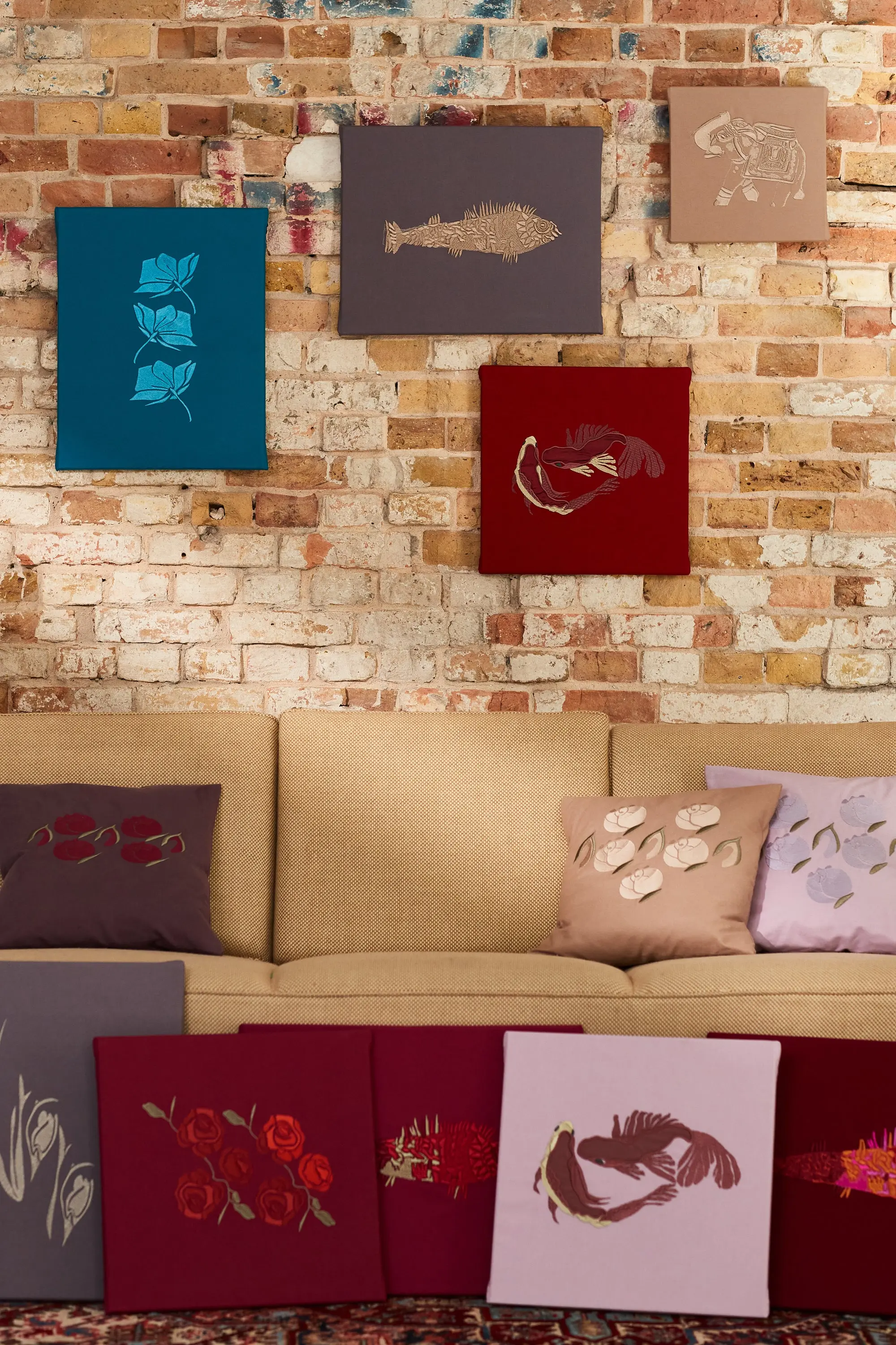 Gestickte Bilder und Kissen in Blau, Rot, Rosa und Beige an einer Backsteinwand und auf einem Sofa